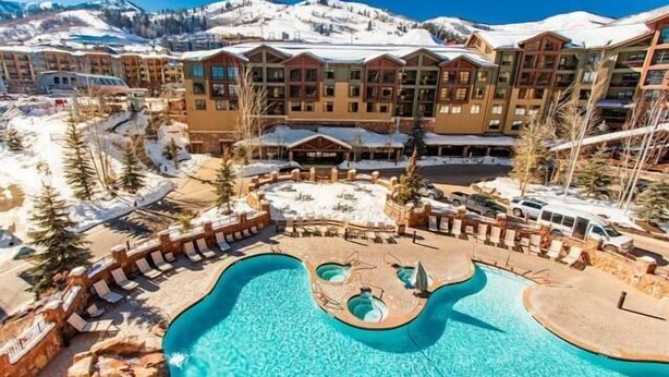 A resort with a large pool, multiple buildings, and snow-covered mountains in the background. - The Grand Summit Hotel in the Canyons Village - Close to Golf - Pool, Gym, Spa, More! (Park City)