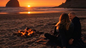 A couple sitting by a campfire on a beach at sunset. - Luxury Beach Home | 1-Min Walk to Sand | 4BR/4BA | Best Location in Shorepine Village (Pacific city)