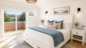 A bedroom with a large bed, a white nightstand, a side table, a view of the outdoors, and a ceiling light fixture. - Luxury Beach Home | 1-Min Walk to Sand | 4BR/4BA | Best Location in Shorepine Village (Pacific city)