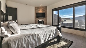 A bedroom with a large bed, a TV mounted on a wooden wall, and a large window with a mountain view.