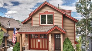 A two-story house with a red garage door, a front porch, and a small front yard with a flag and a mailbox. - Beautiful Historic Town Home, Walk to Main St - Large Deck w/ Mountain Views and Private Hot Tub! (Park City)