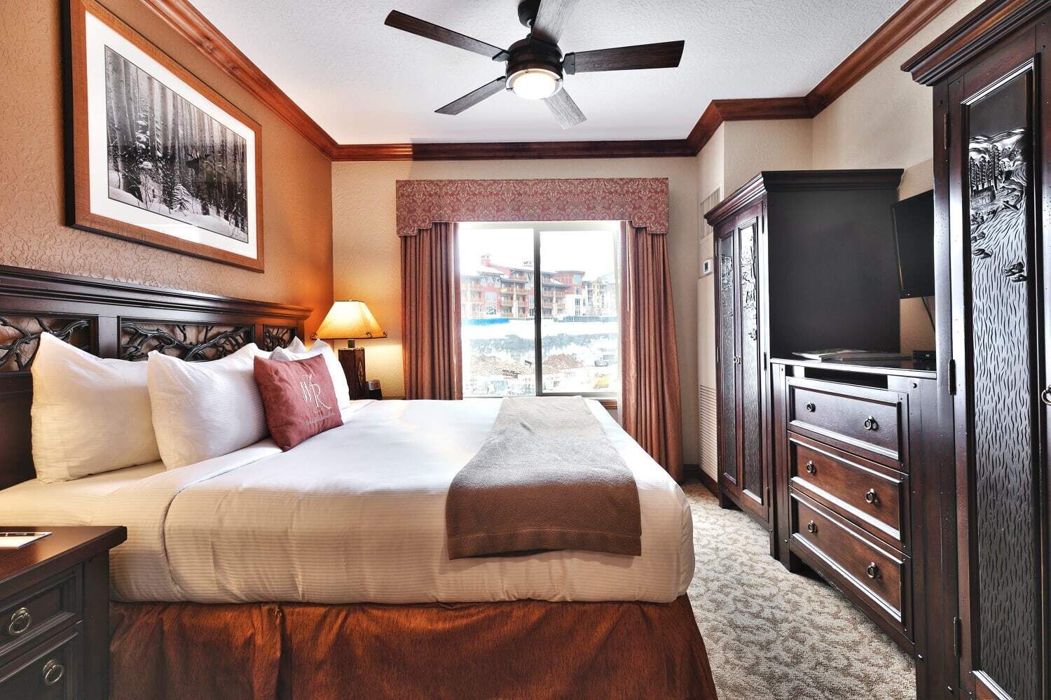 A hotel room with a large bed, a ceiling fan, a TV, and a view of the city through the window.