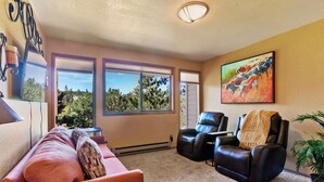 A living room with a sofa, two leather chairs, a painting, and a view through large windows. - Multiple Decks + Solarium Sitting Room, Office Space & Dog Friendly! (Woodland Park)