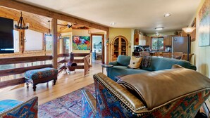 A spacious living room with a large sofa, a patterned armchair, and a wooden coffee table. There is a dining area with a kitchen visible in the background. - Multiple Decks + Solarium Sitting Room, Office Space & Dog Friendly! (Woodland Park)