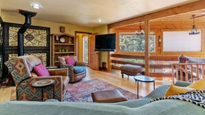 A cozy living room with a fireplace, a television, and a large window overlooking a forest. - Multiple Decks + Solarium Sitting Room, Office Space & Dog Friendly! (Woodland Park)