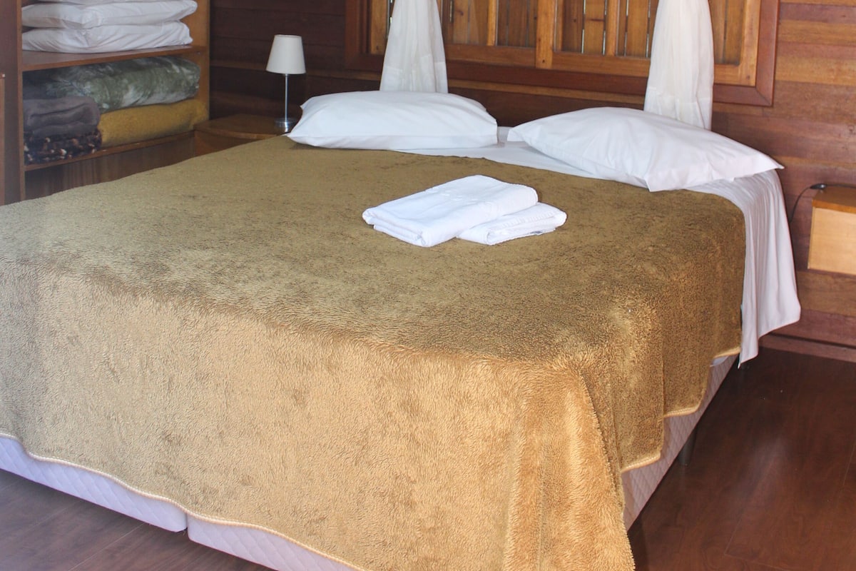 Una cama bien hecha con una colcha marrón, almohadas blancas y un dosel blanco. Se observa un armario de madera con toallas dobladas y una barra para colgar ropa.