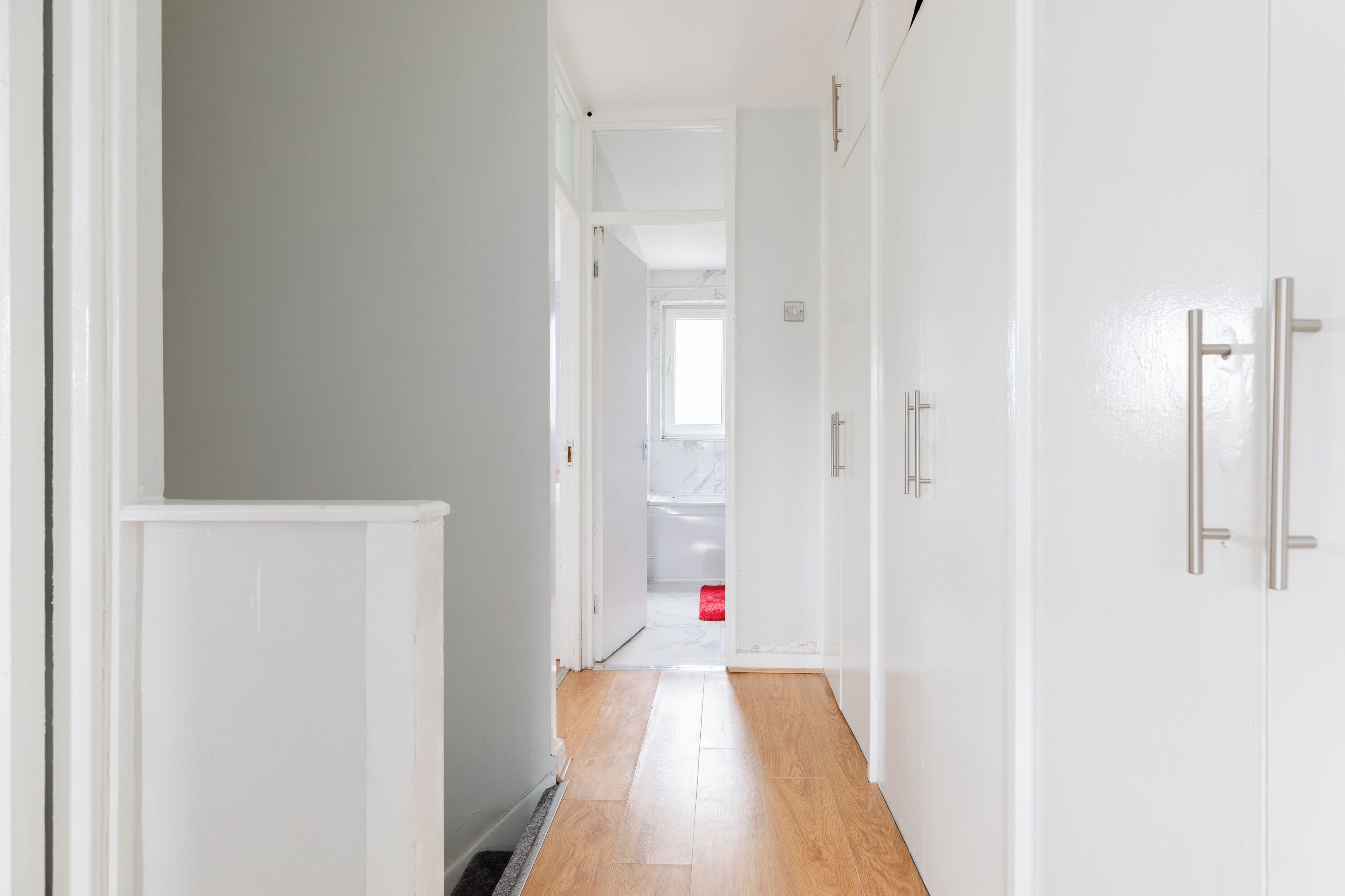 Un pasillo con paredes blancas, piso de madera y una puerta que da al baño.