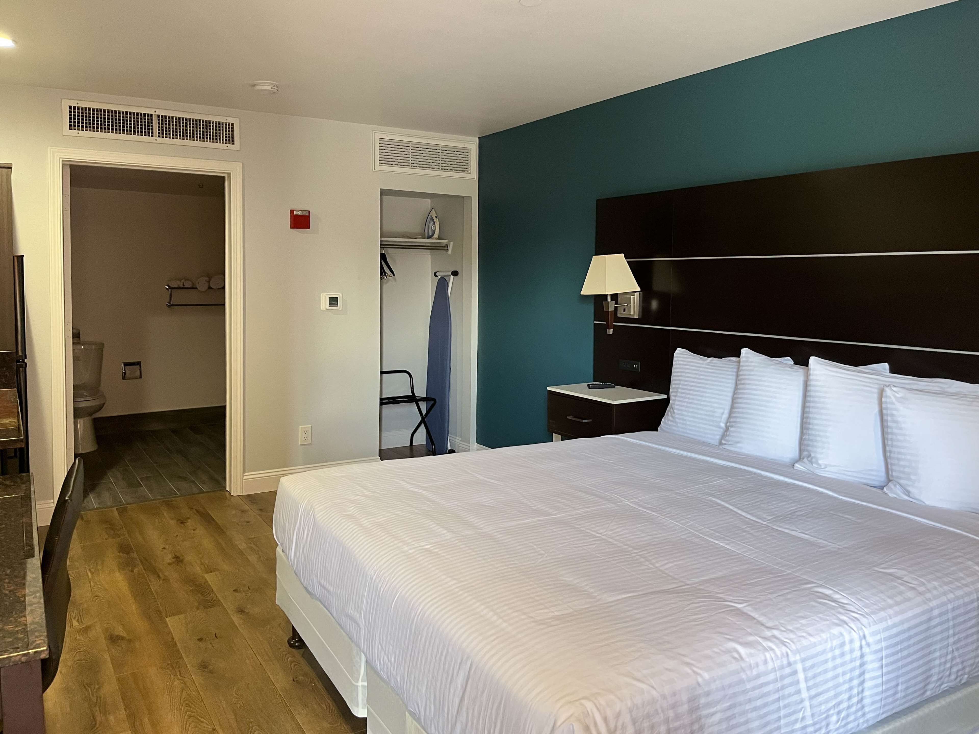 A hotel room with a large bed, a nightstand with a lamp, a wardrobe, and a bathroom visible through an open door.