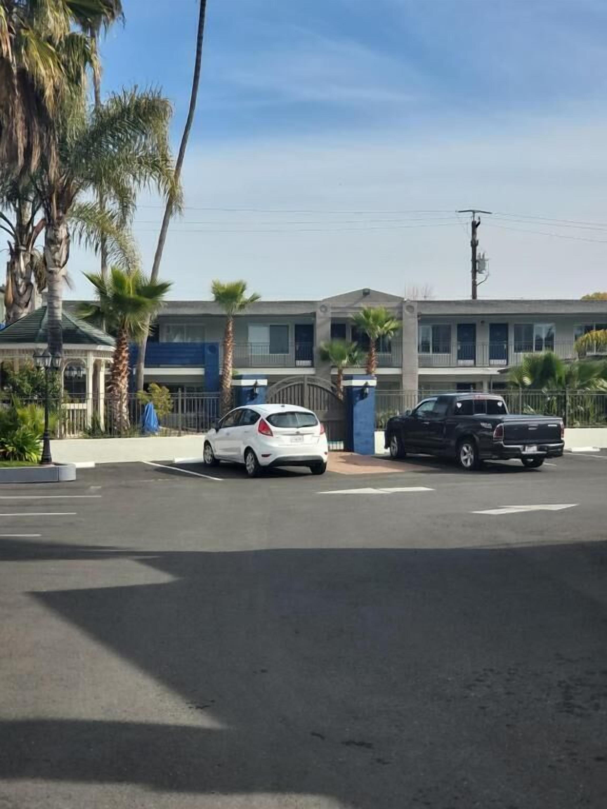 A parking lot with a white car and a black truck, a building with a blue facade, and palm trees.