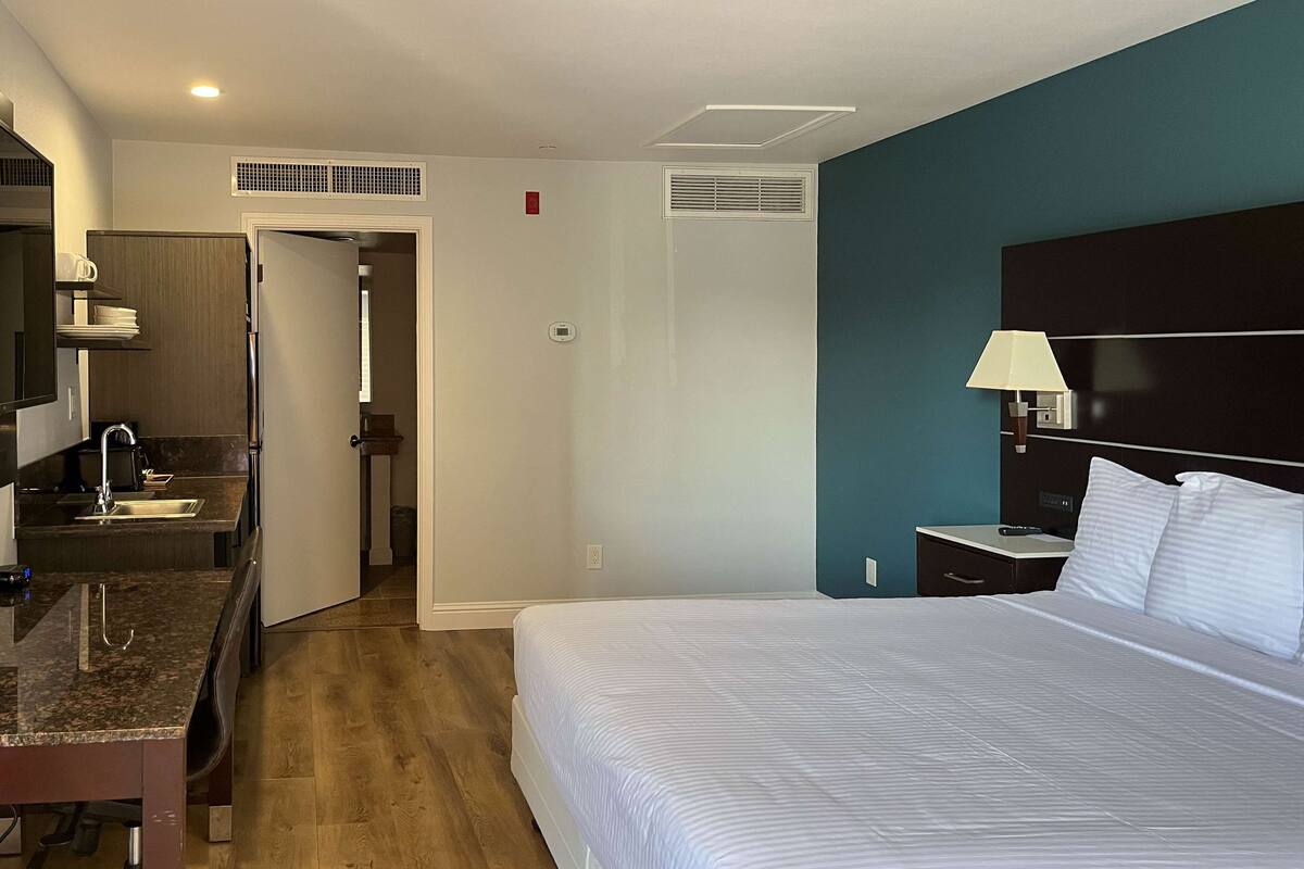 A hotel room with a bed, a desk, a chair, a nightstand, a lamp, and a bathroom visible through an open door.