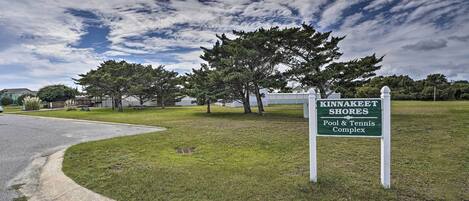 A sign for Kinnakeet Shores Pool & Tennis Complex with a grassy area and trees in the background.