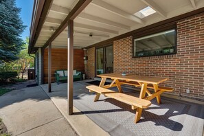 Ein überdachter Patio-Bereich mit Holztisch und Bänken, einer Ziegelwand und einem großen Fenster.
