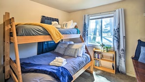 Een slaapkamer met een stapelbed, een raam, een klein houten plankje met boeken en een plant.
