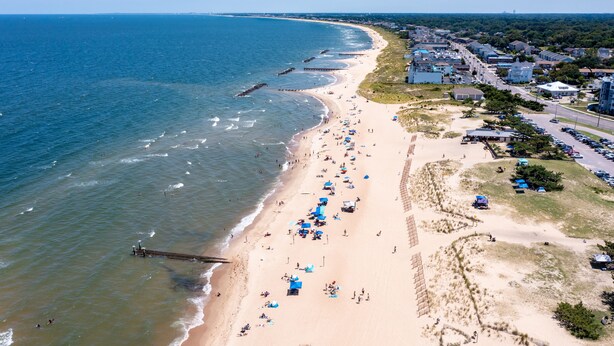Ein Strand mit einem Steg, der ins Wasser führt, ein sandiger Uferbereich mit Sonnenschirmen und eine Wohngegend mit Gebäuden und Straßen in der Nähe.