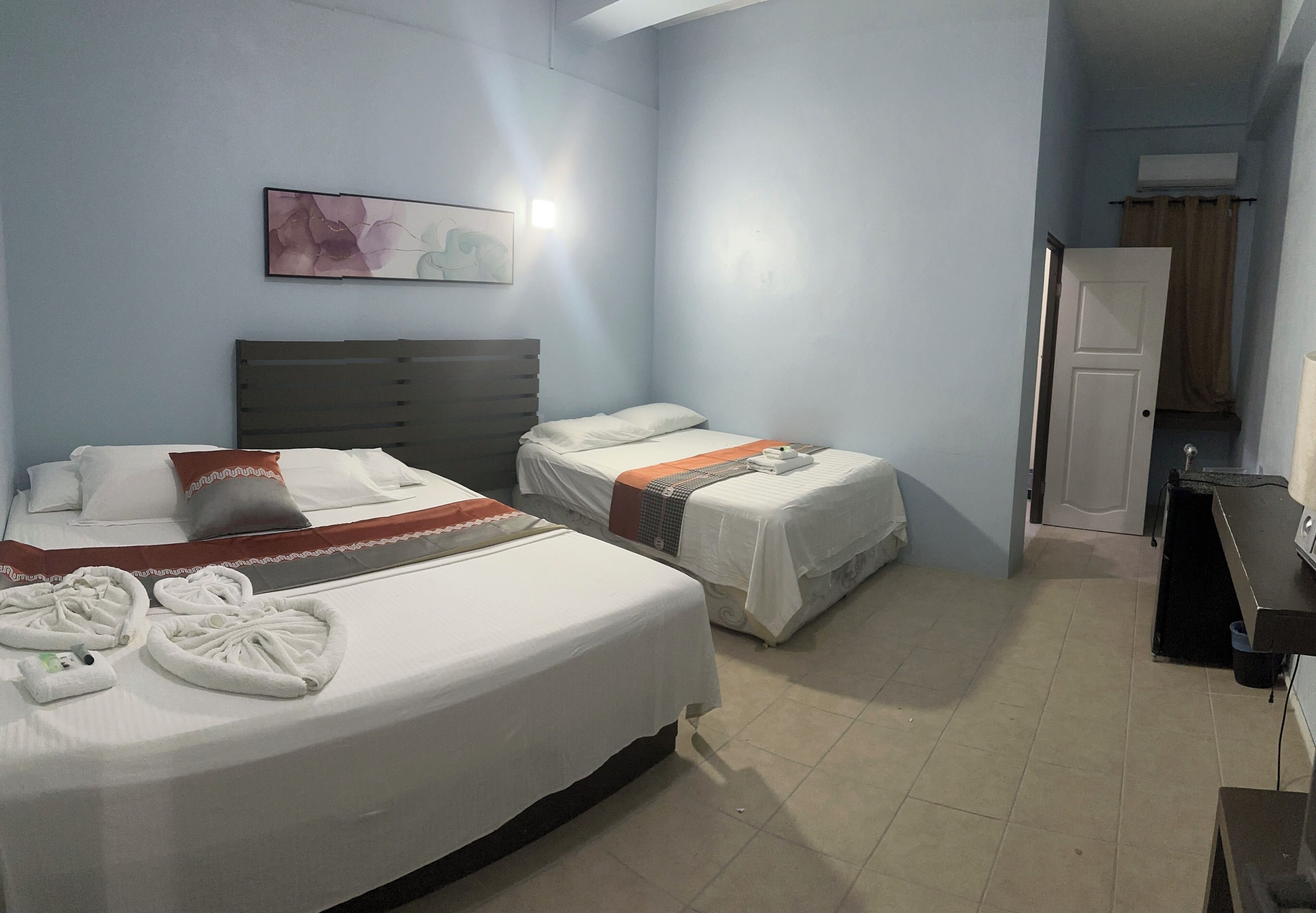 A hotel room with two beds, a desk, a chair, a painting on the wall, and a door leading to a bathroom.