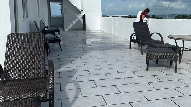 A rooftop terrace with wicker furniture, a staircase leading to the upper level, and a view of the sea under a cloudy sky.