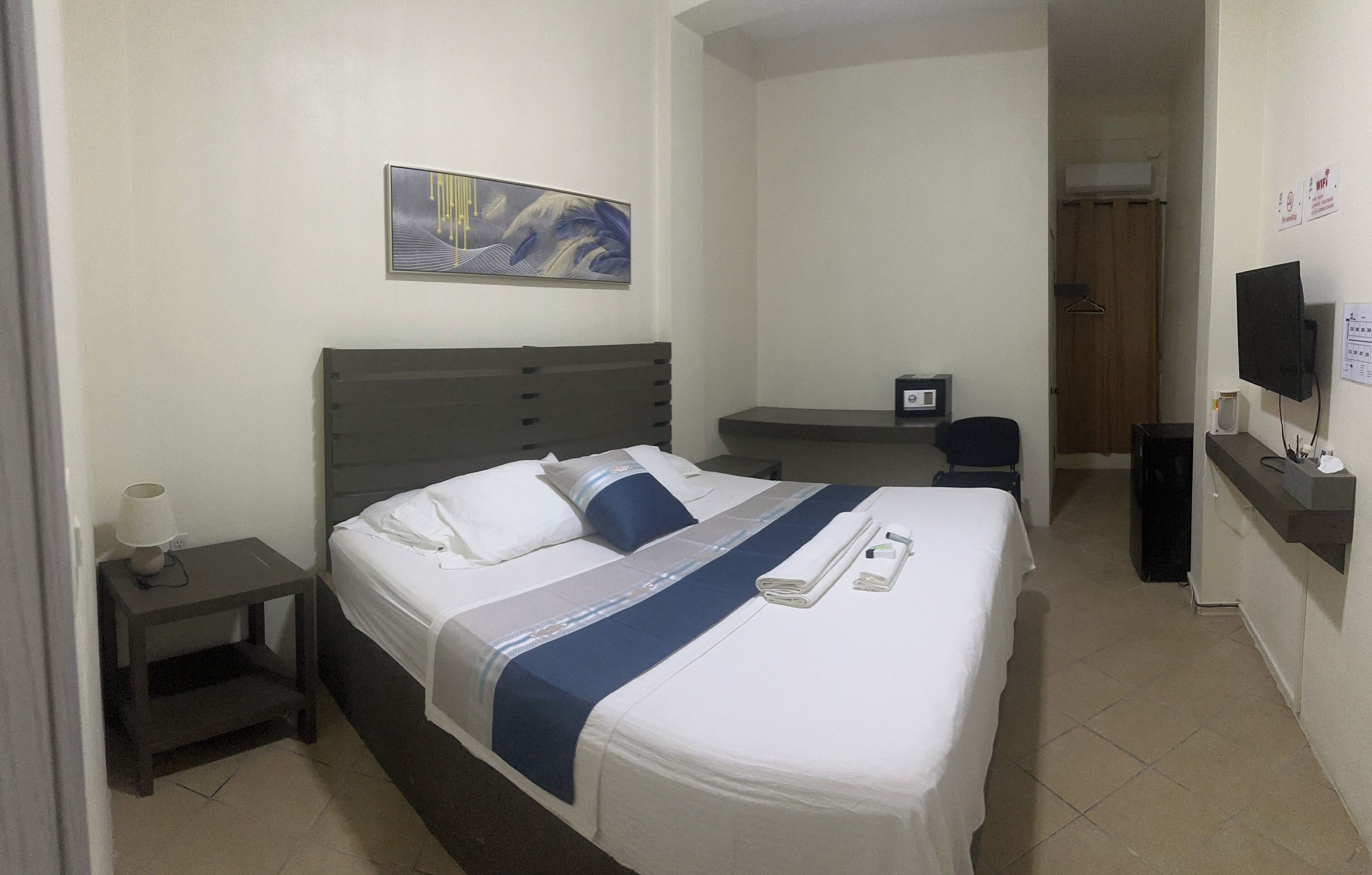 A hotel room with a bed, bedside table, lamp, artwork, TV, and a desk with a chair.