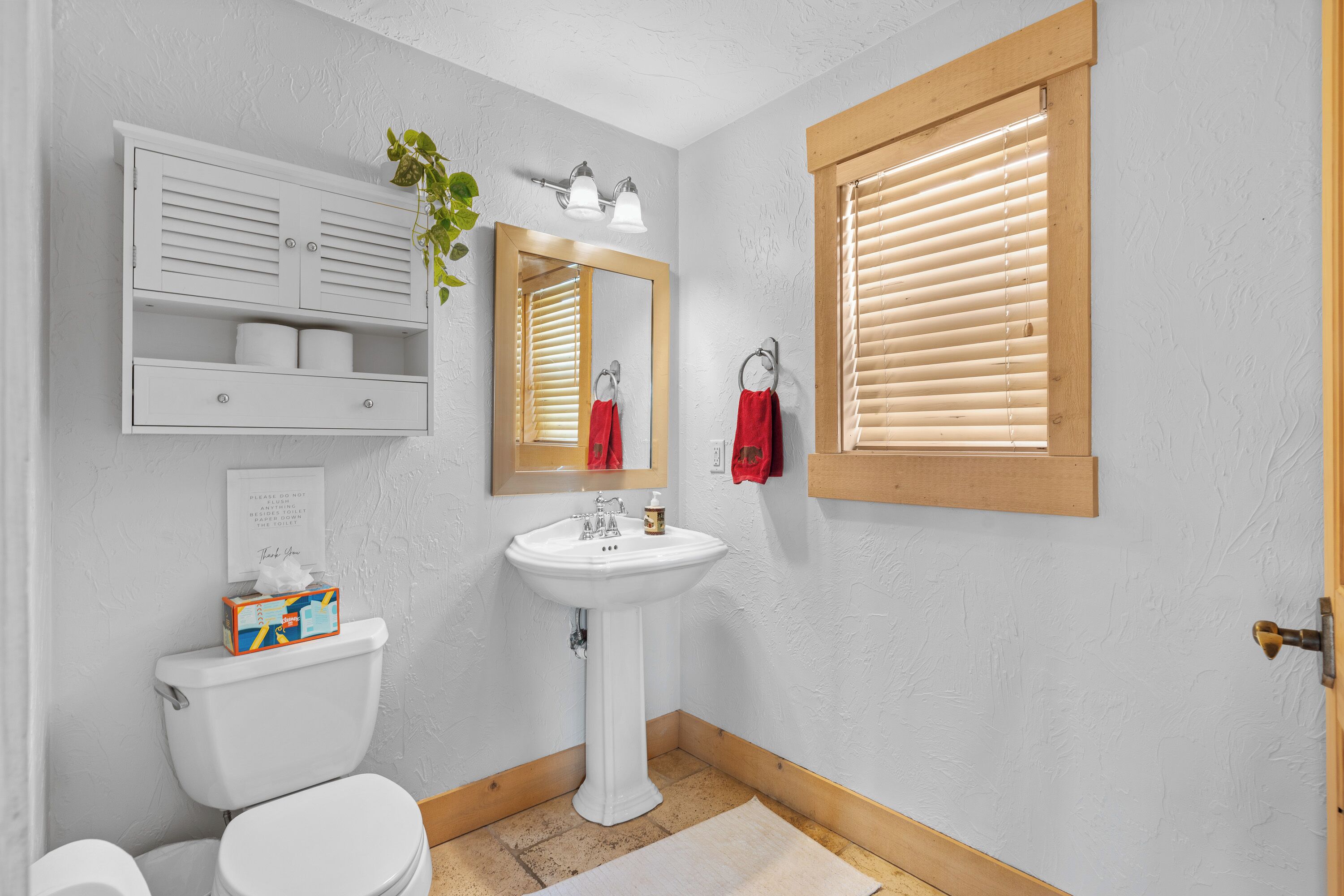 A bathroom with a white pedestal sink, a toilet, a mirror, a wooden window blind, and a shelf with toiletries.