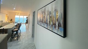 A modern living room with a large abstract painting, a dining area with bar stools, and a view of the sea through large windows.