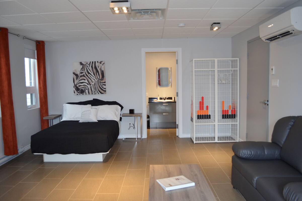 A modern bedroom with a bed, bedside table, and a view into the kitchen.