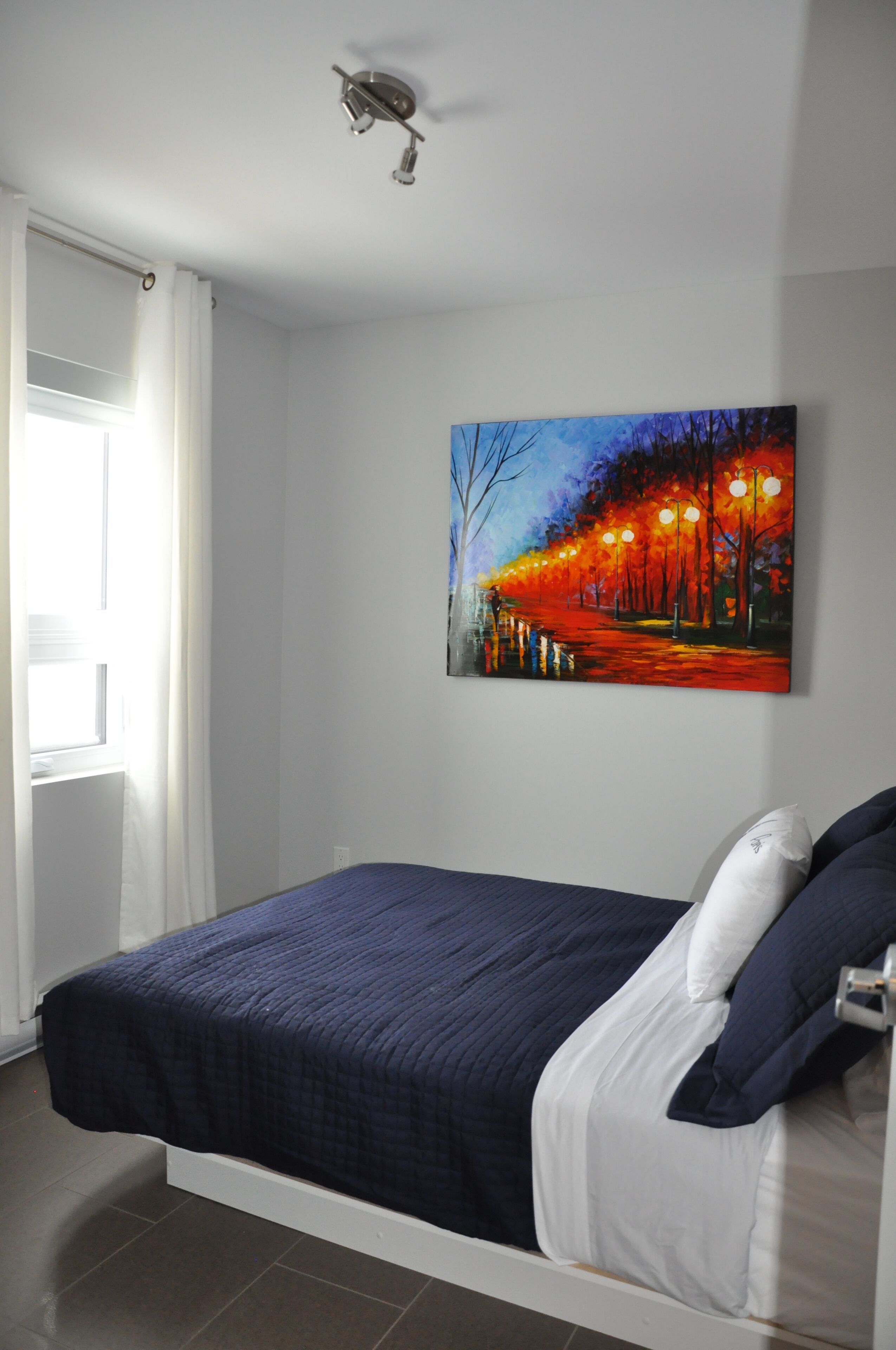 A bedroom with a bed, a painting of a street scene, and a window with curtains.