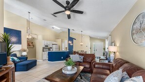 A spacious living room with a brown leather sofa, a blue armchair, and a wooden coffee table. A ceiling fan and pendant lights are visible. - Vibrant resort pool home with games room - Terra Verde (Kissimmee)