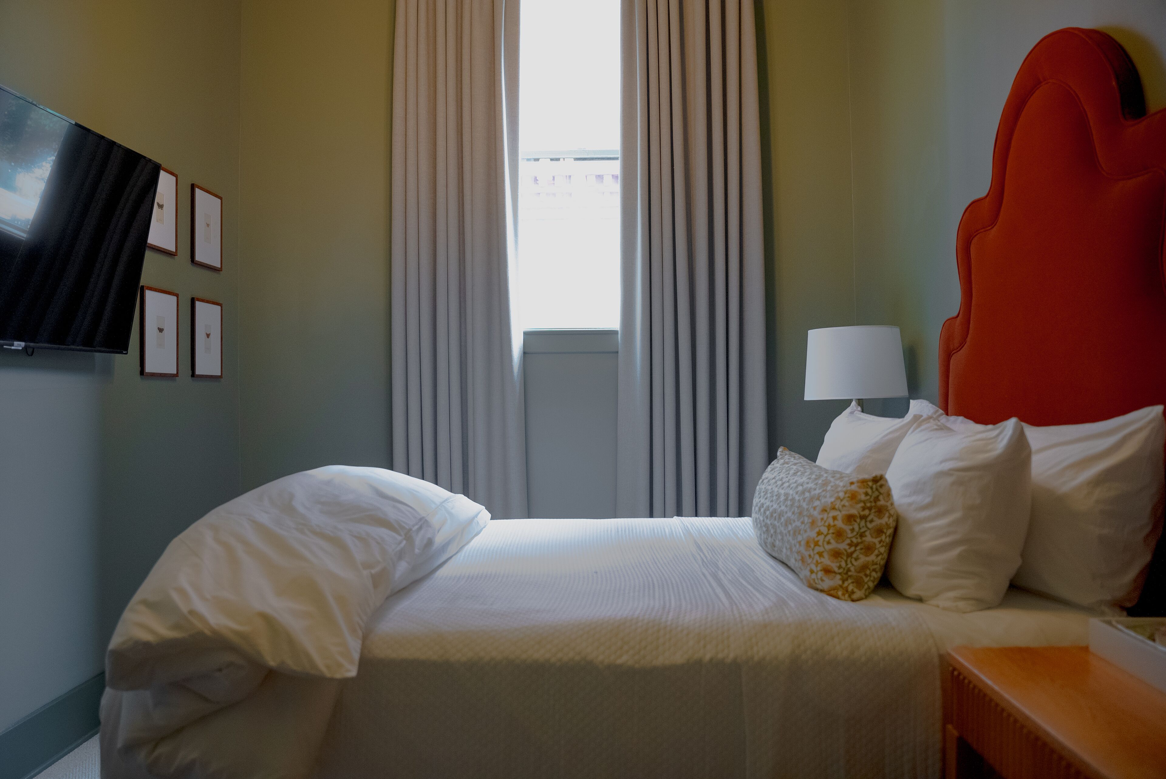 A hotel room with a bed, a TV mounted on the wall, a window with curtains, and a red headboard.