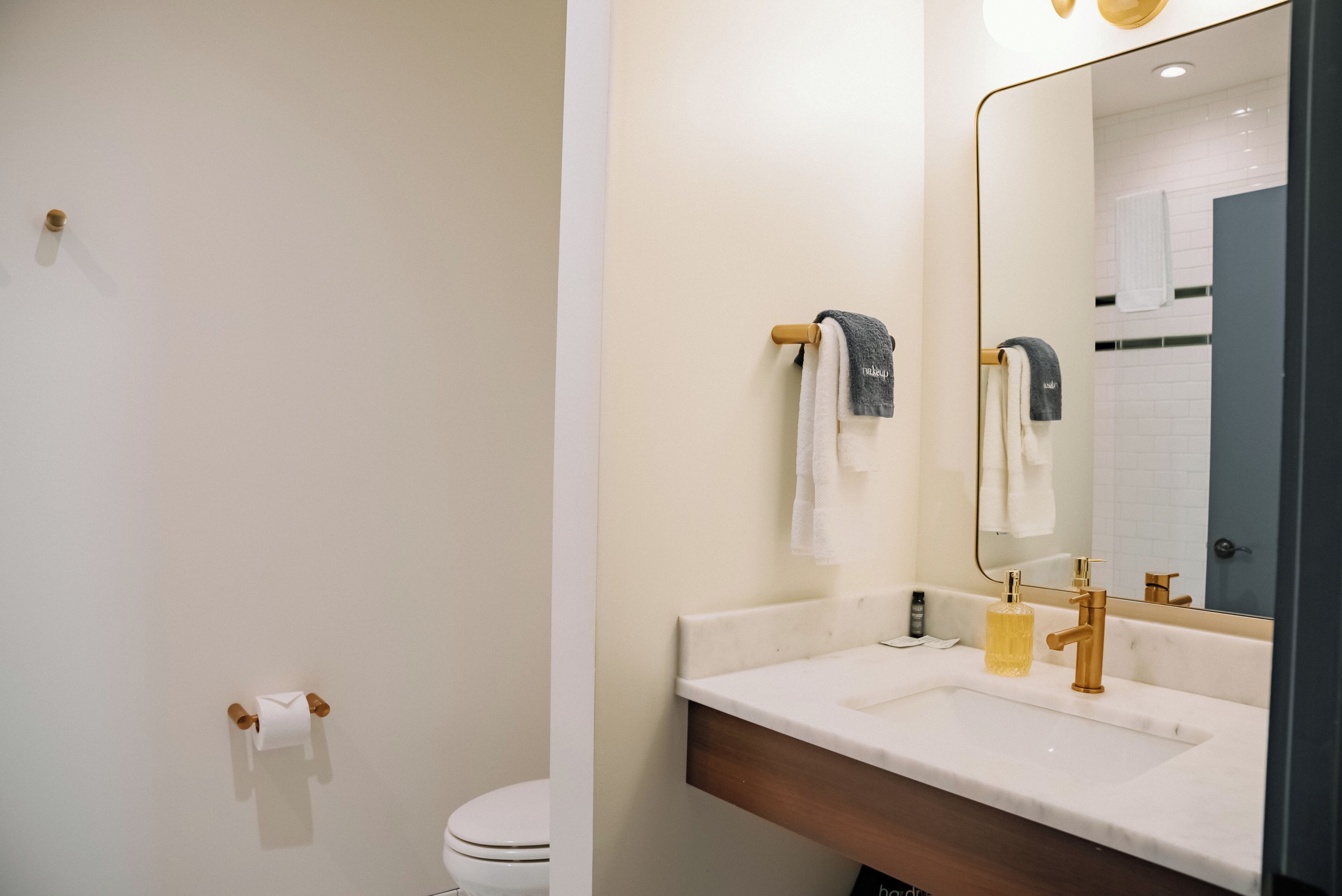 Ein modernes Badezimmer mit einem weißen Waschbecken, einem goldenen Wasserhahn und einem großen Spiegel. Handtücher hängen an einem hölzernen Handtuchhalter und ein Toilettenpapierhalter ist an der Wand angebracht.