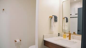 A modern bathroom with a white sink, gold faucet, and a large mirror. There are towels hanging on a wooden towel rack and a toilet paper holder on the wall.
