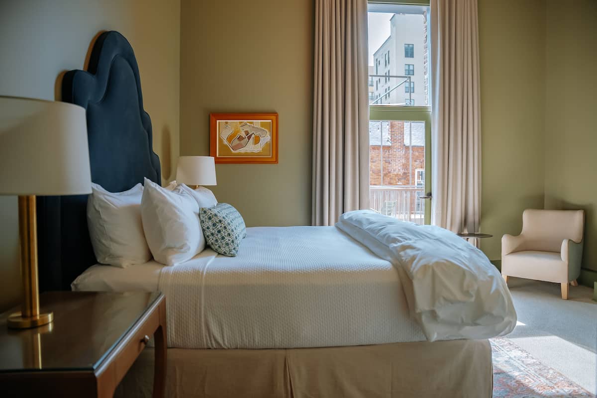 A hotel room with a bed, two bedside tables, a lamp, a chair, a window with curtains, and a framed picture on the wall.