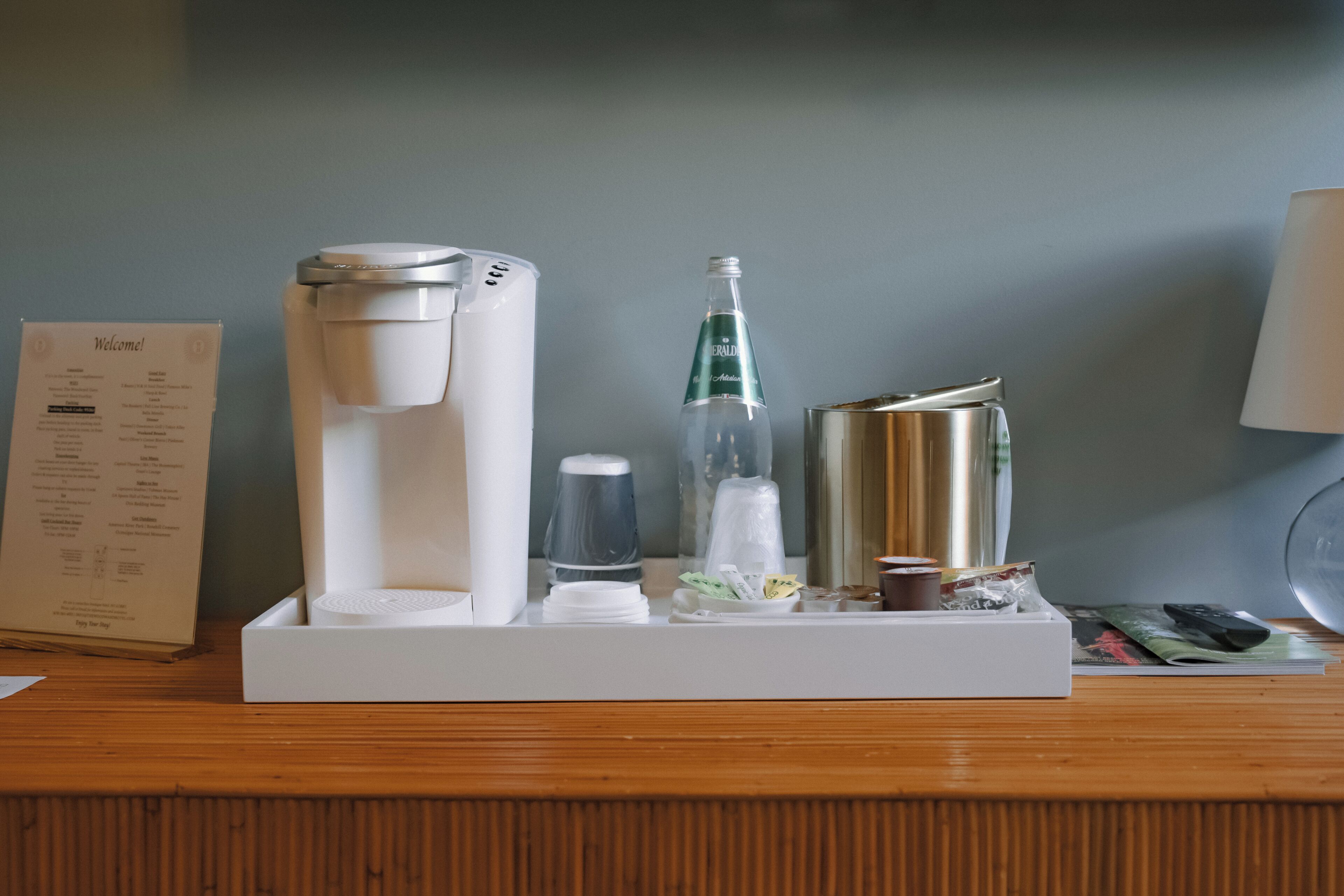De receptie van een hotelkamer met een koffiezetapparaat, een waterfles, een glas, een klein bakje en een menukaart op een houten tafel.