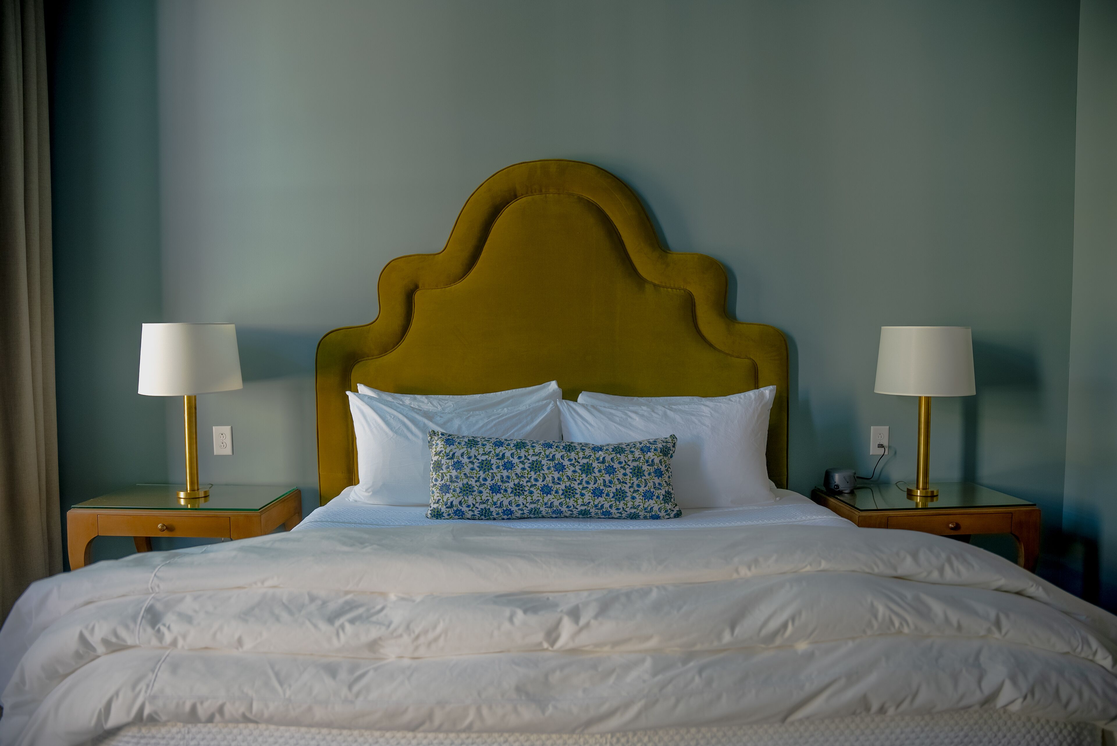 A neatly made bed with a wooden headboard, white bedding, and a patterned pillow. There are two bedside tables with lamps and a small drawer.