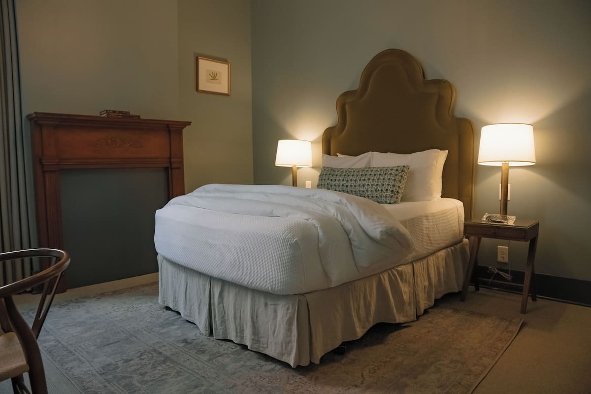 A neatly made bed with a wooden headboard, two bedside lamps, and a chair in a room with a fireplace and a framed picture on the wall.