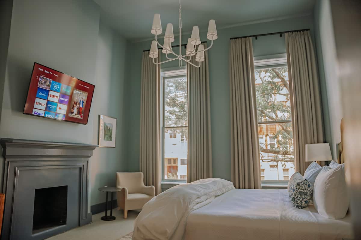 A hotel room with a fireplace, a large bed, a TV mounted on the wall, and a view of the outside through the window.