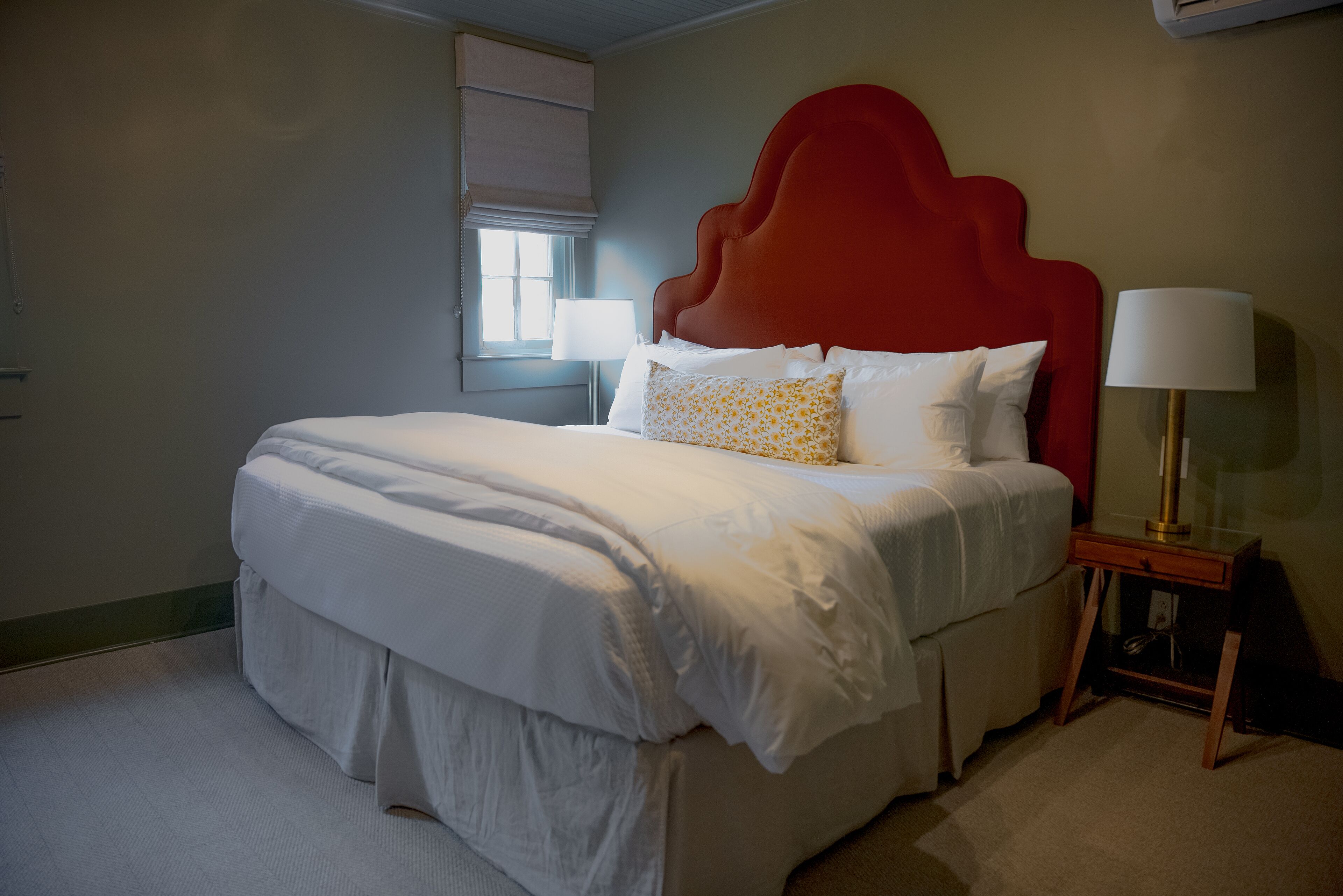 Een net opgemaakt bed met witte lakens, een rood hoofdeinde en een bedlampje.