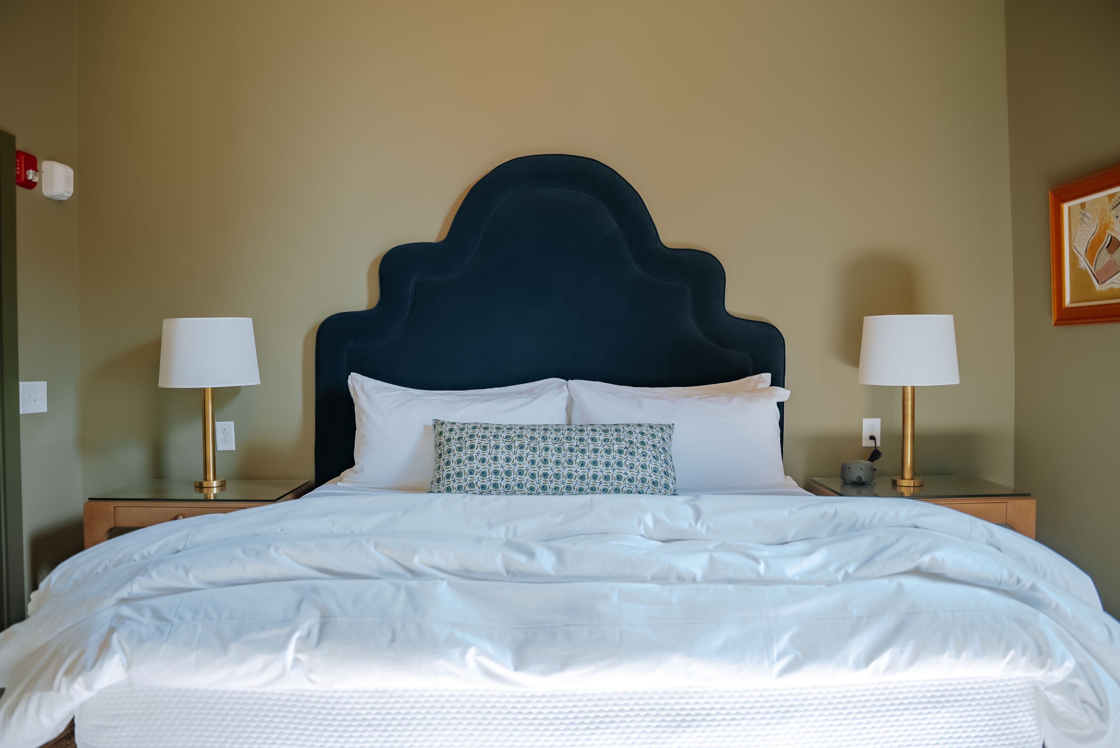 Una cama bien tendida con sábanas blancas, un cabecero tapizado en azul oscuro y dos lámparas de noche con pantallas blancas.