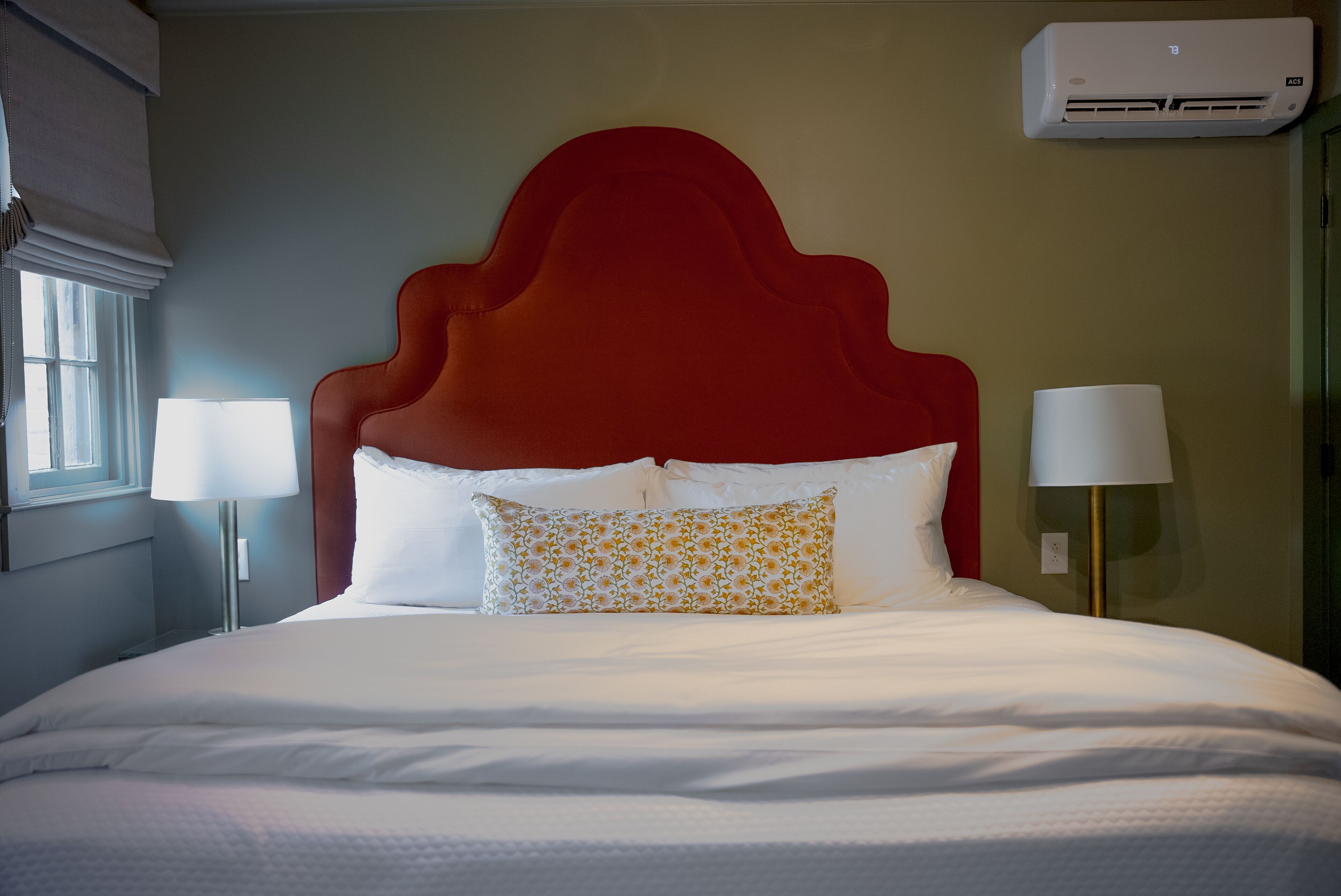 A hotel room with a large bed, two bedside lamps, a red headboard, and an air conditioning unit.