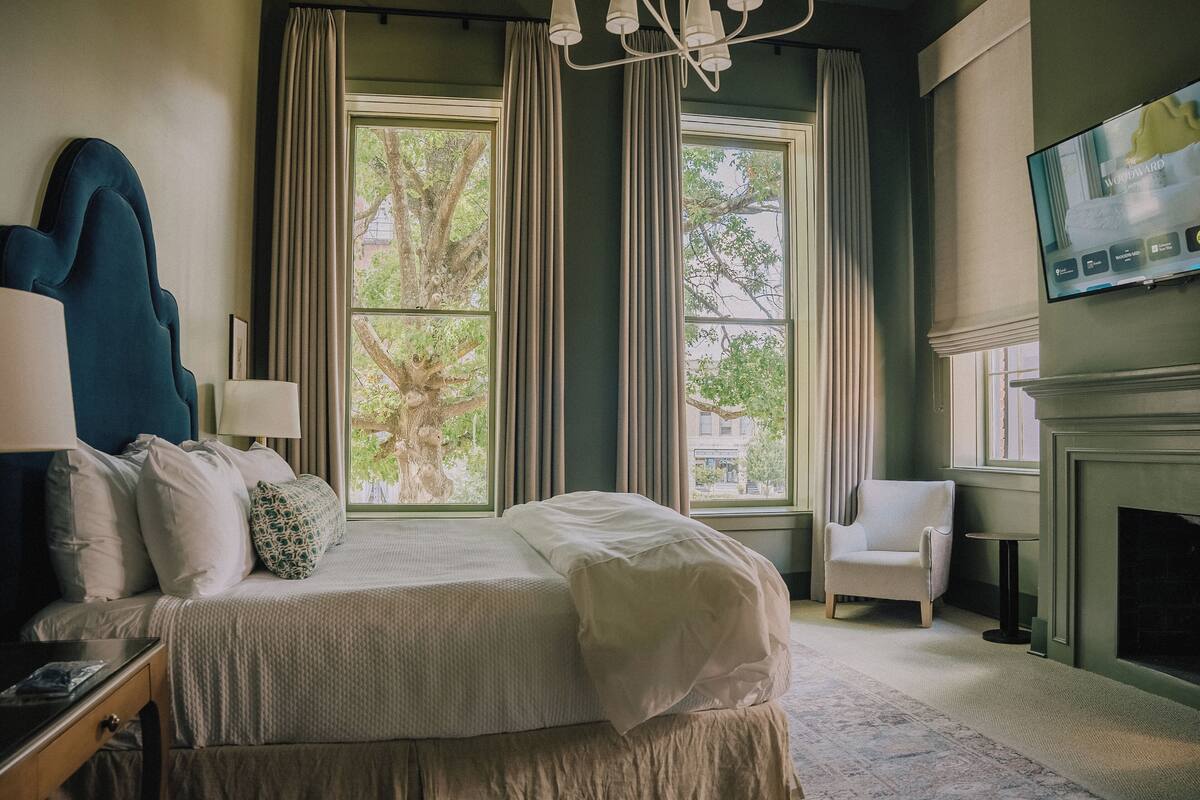 A neatly arranged bedroom with a bed, bedside lamps, a chair, a fireplace, and a TV mounted on the wall.