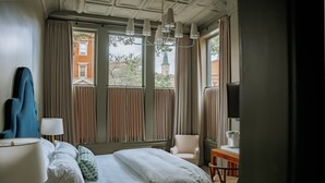 A hotel room with a bed, a chair, a table, a lamp, and a view of a building with a green roof. - The Woodward Hotel (Macon)