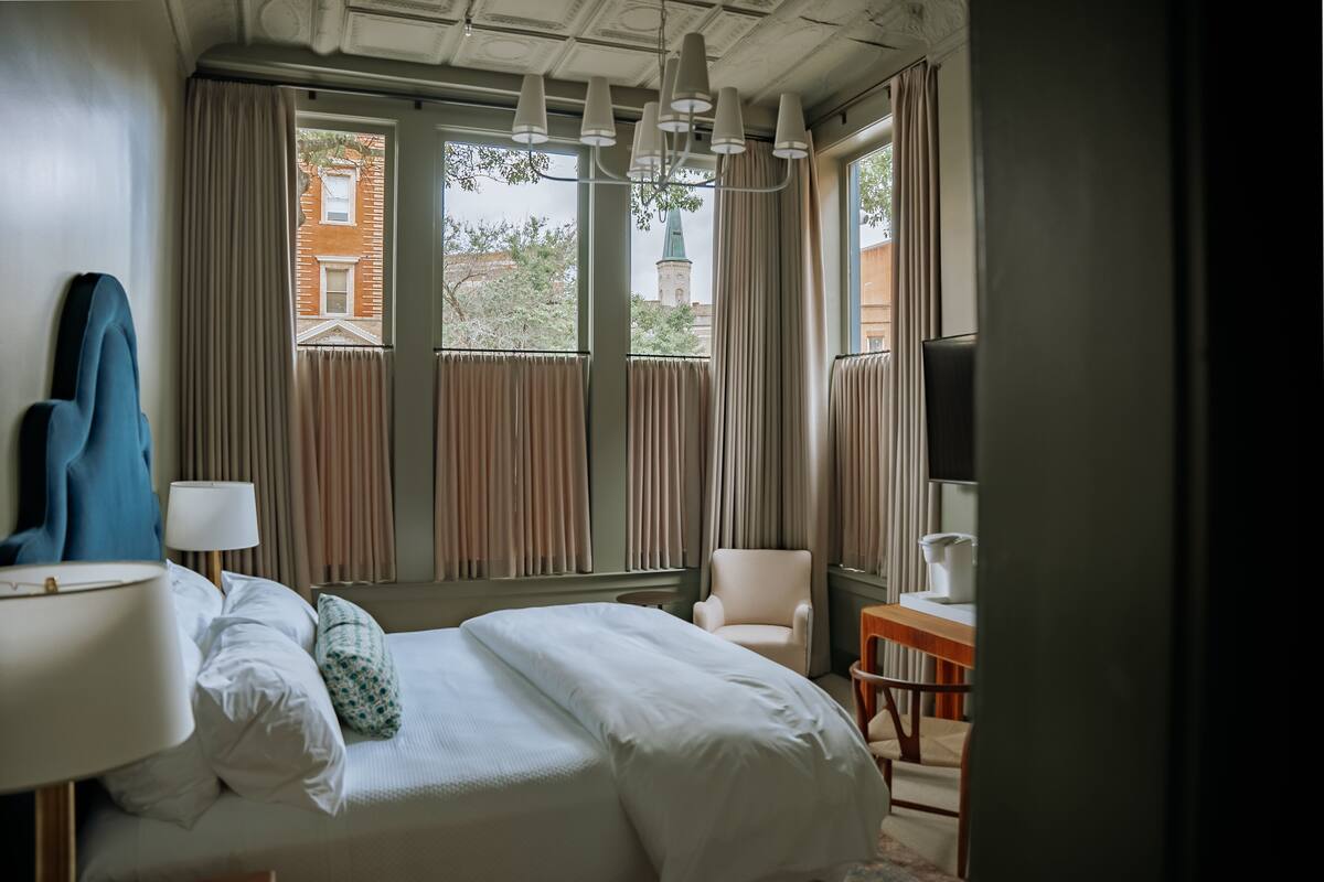 A hotel room with a bed, a chair, a table, a lamp, and a view of a building with a green roof.