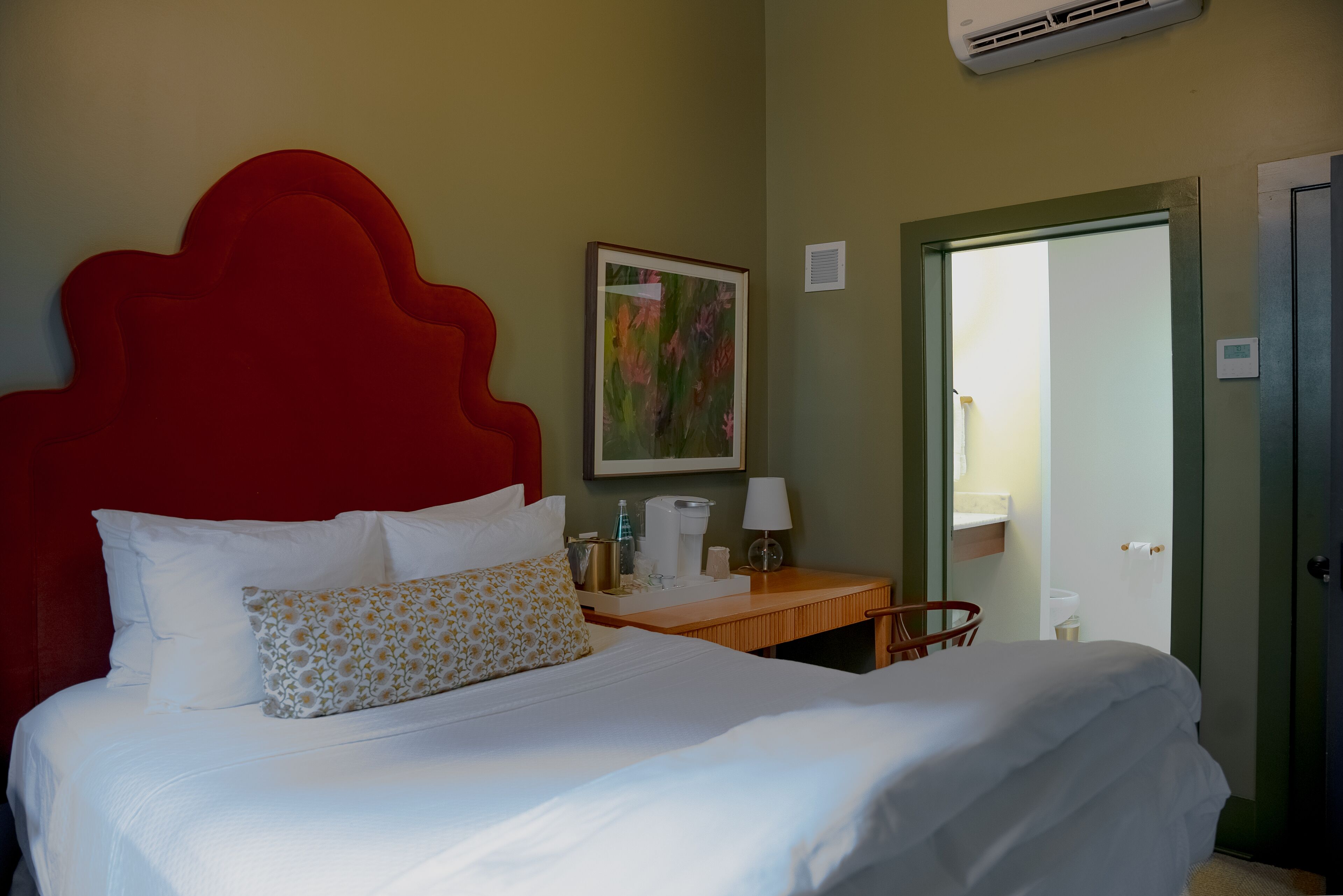 A hotel room with a large bed, a red headboard, a bedside table with a lamp, a painting on the wall, and a door to the right.