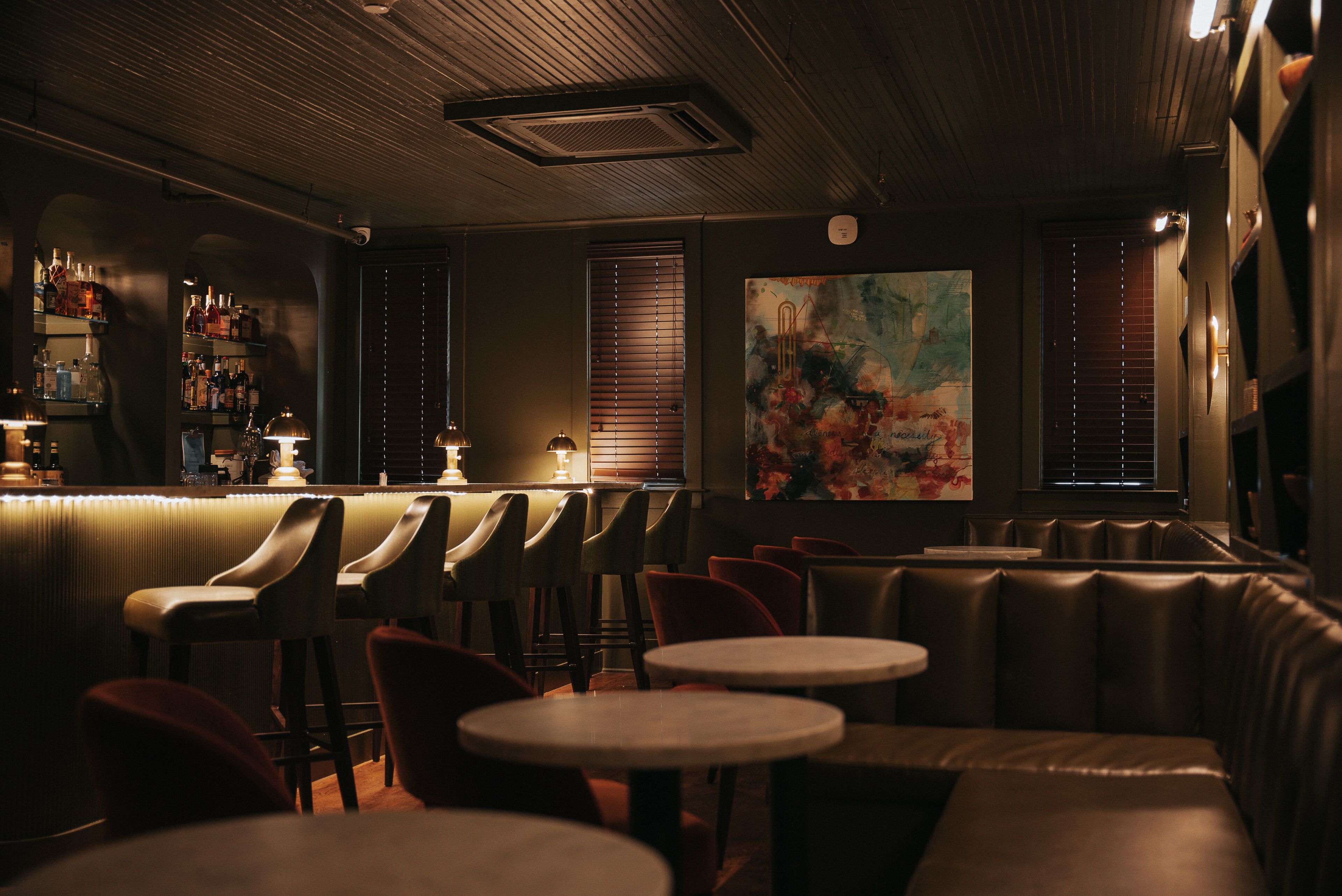 A dimly lit bar area with leather booths and round tables, a bar counter with bottles, and a large abstract painting on the wall.