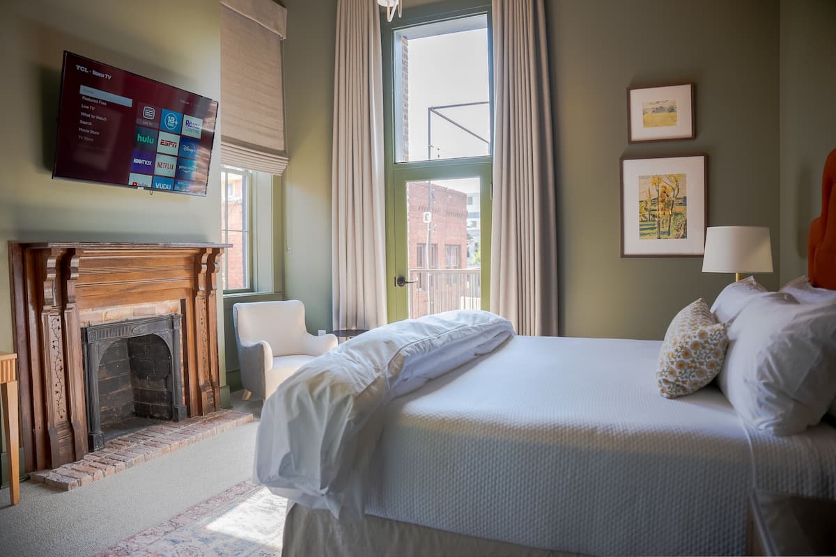 A hotel room with a bed, a fireplace, a TV mounted on the wall, and a window with curtains.