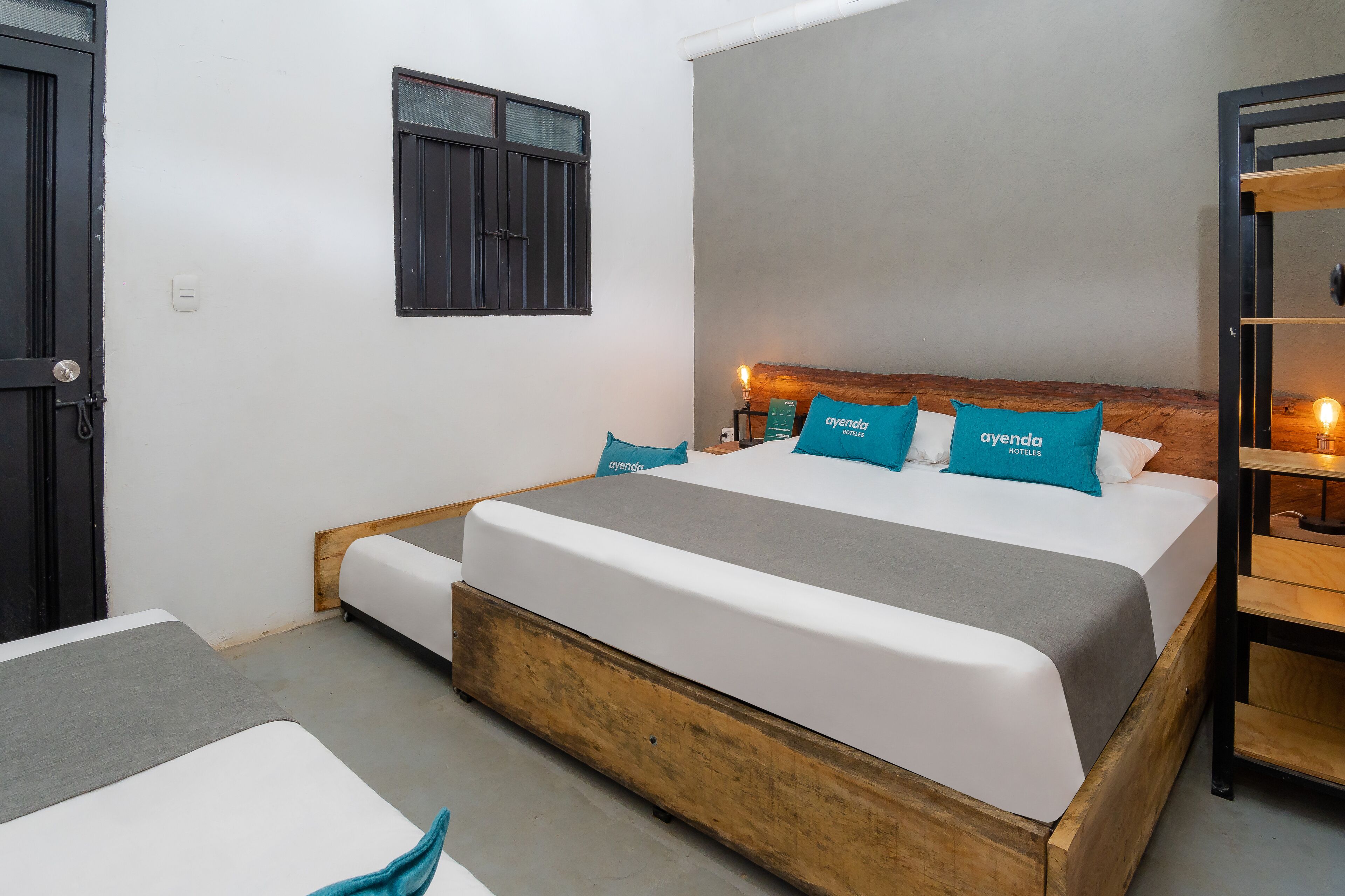 Un letto singolo con struttura in legno, biancheria da letto bianca e cuscini color petrolio con la scritta "ayenda HOTELS".