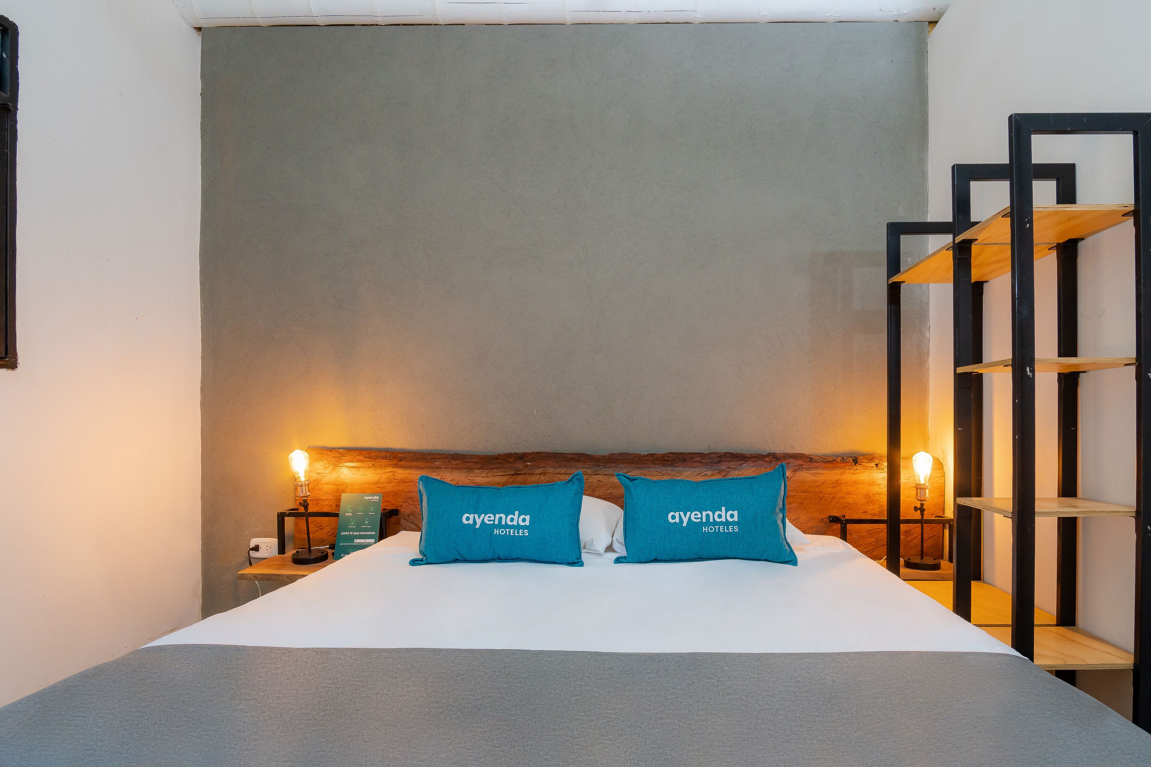 A hotel room with a bed, two pillows with the hotel's name, a nightstand with a lamp, and a wooden shelf unit.