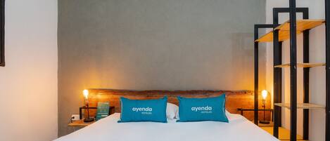 A hotel room with a bed, two pillows with the hotel's name, a nightstand with a lamp, and a wooden shelf unit.