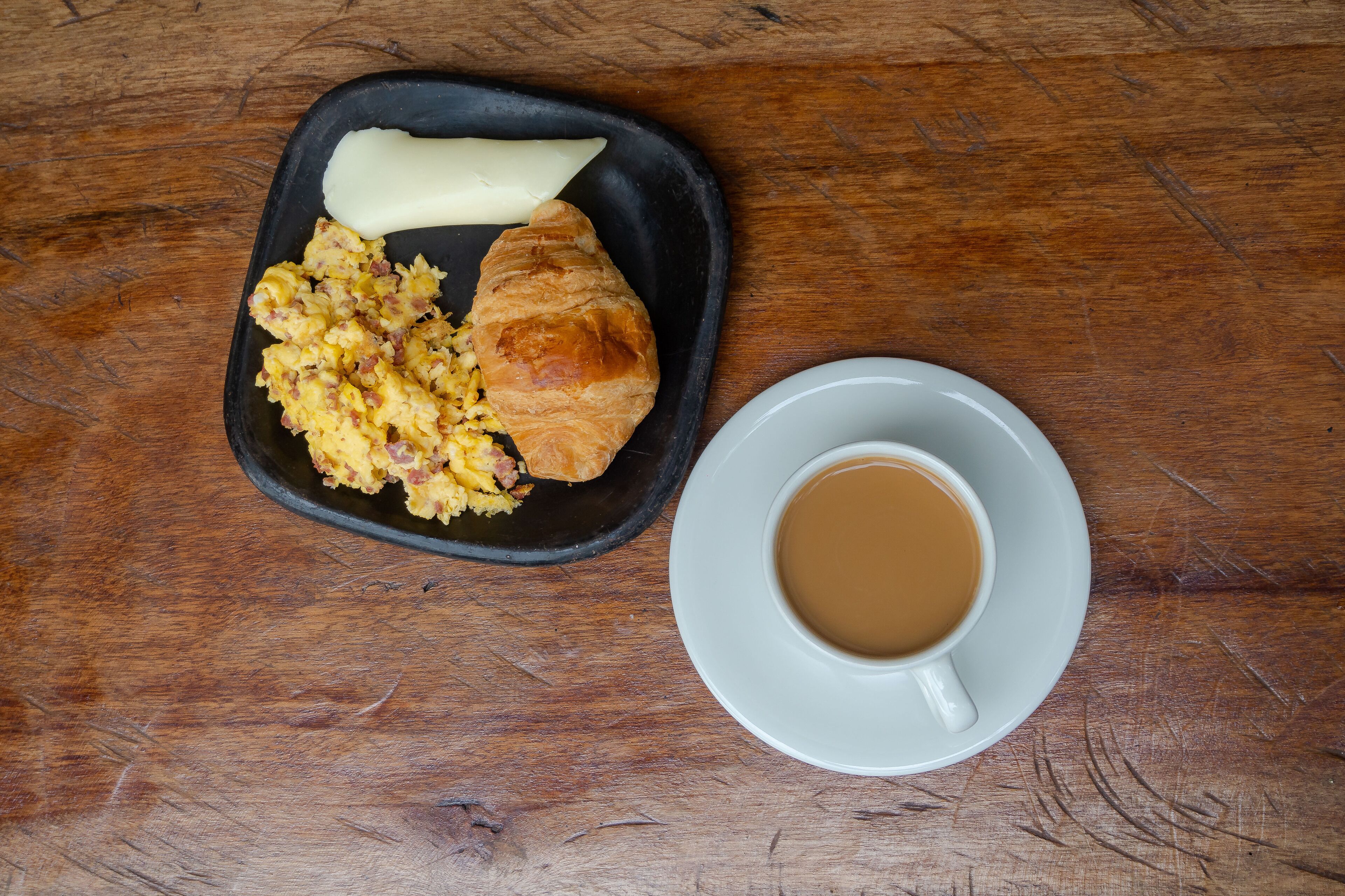 Um prato preto com ovos mexidos, um croissant e uma porção de manteiga sobre uma mesa de madeira. Ao lado, uma chávena branca com pires contém um líquido castanho, provavelmente café.
