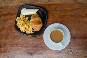 Um prato preto com ovos mexidos, um croissant e uma porção de manteiga sobre uma mesa de madeira. Ao lado, uma chávena branca com pires contém um líquido castanho, provavelmente café.