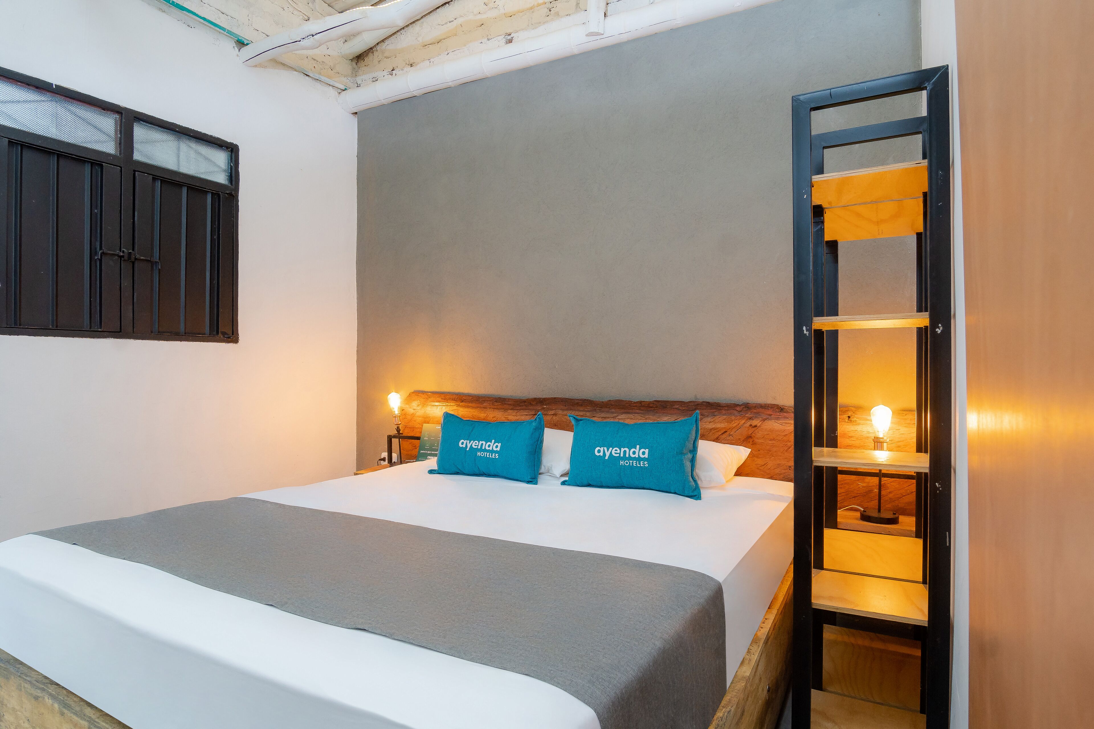 A neatly made bed with a grey bedspread and two blue pillows, a wooden headboard, a wall-mounted shelf with a light, and a door with a wooden frame.