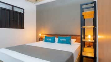 A neatly made bed with a grey bedspread and two blue pillows, a wooden headboard, a wall-mounted shelf with a light, and a door with a wooden frame.